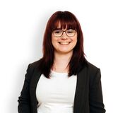 A smiling woman in glasses stands confidently, wearing a black blazer and jeans against a white background.
