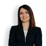 A confident woman stands with arms crossed, wearing a black blazer and jeans against a white background.