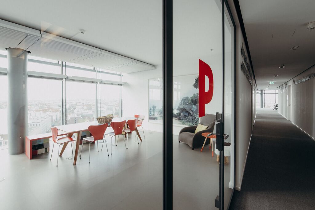 A modern office space with red chairs and a large window overlooks the city, while a hallway stretches into the distance.